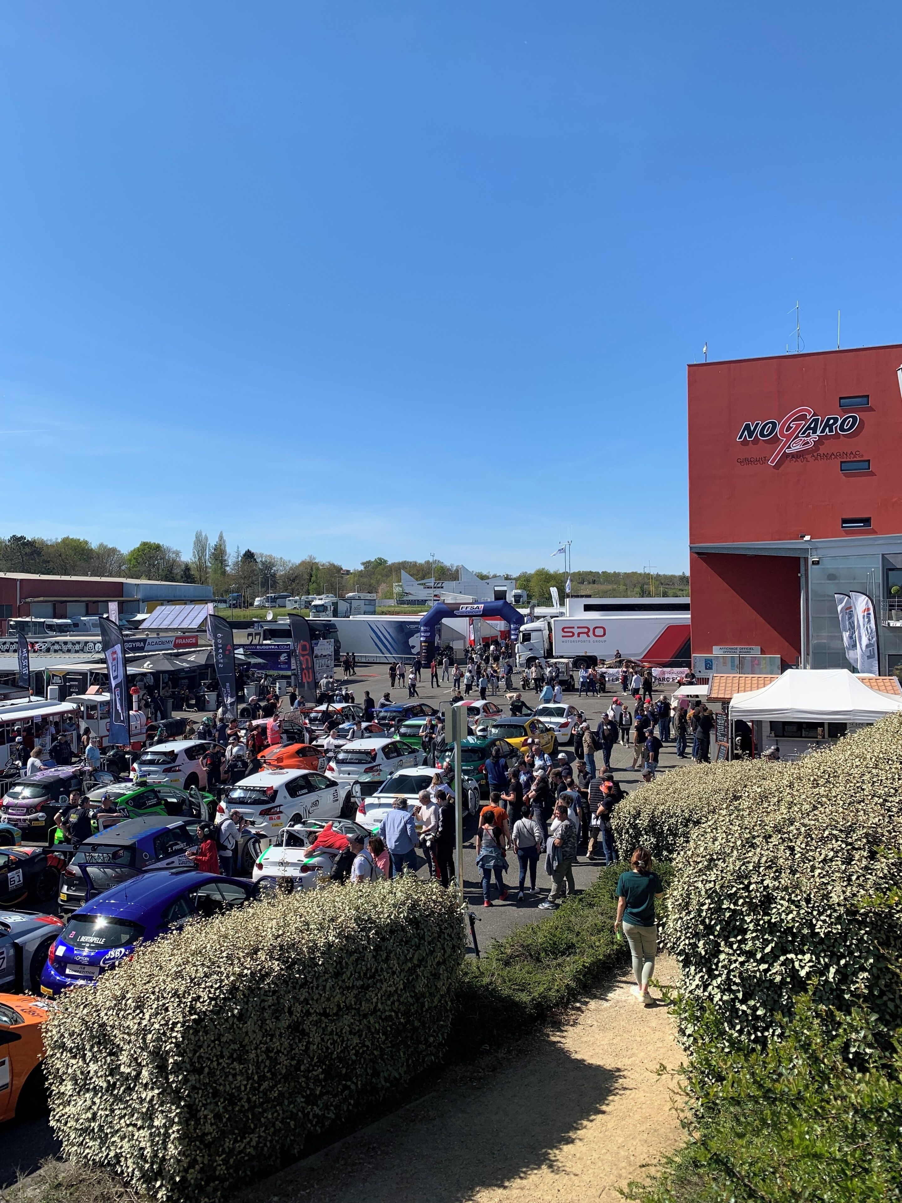 A busy outdoor event with numerous cars and people, tents, and a large red building with signage.