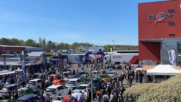 A busy outdoor event with numerous cars and people, tents, and a large red building with signage.