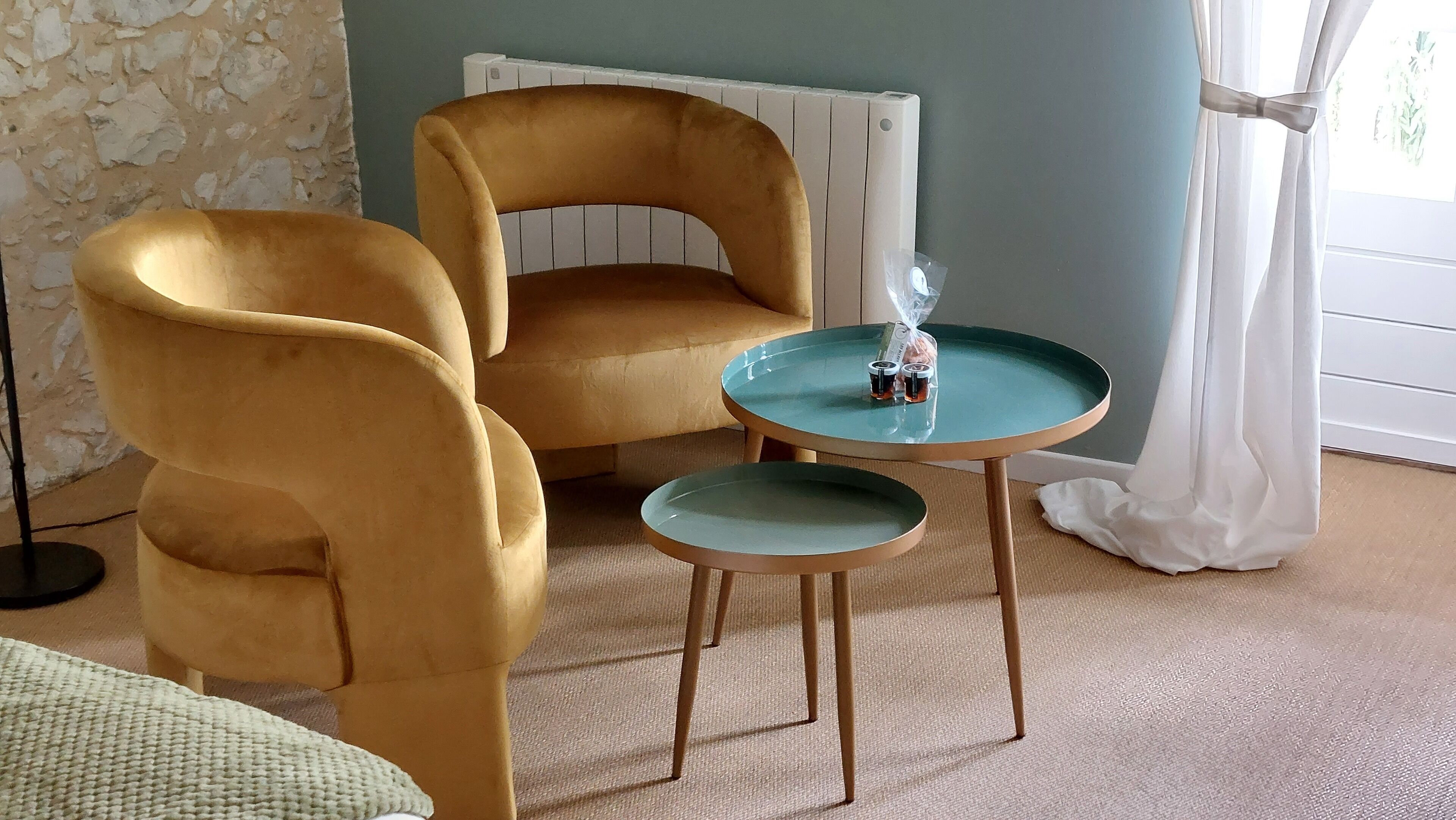 A room with two mustard-colored armchairs, a small teal table with a bottle and a container, a radiator, and a window with white curtains.