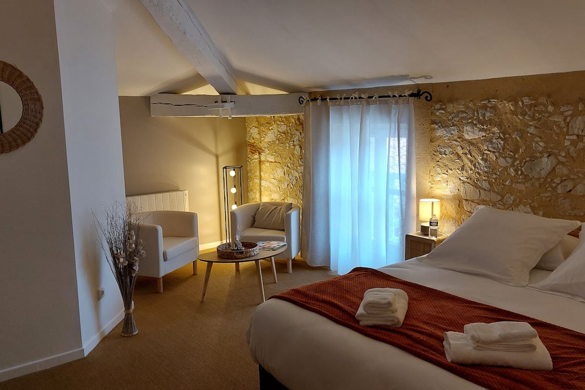 A hotel room with a bed, a seating area with a chair and a table, a stone wall, and a window with curtains.