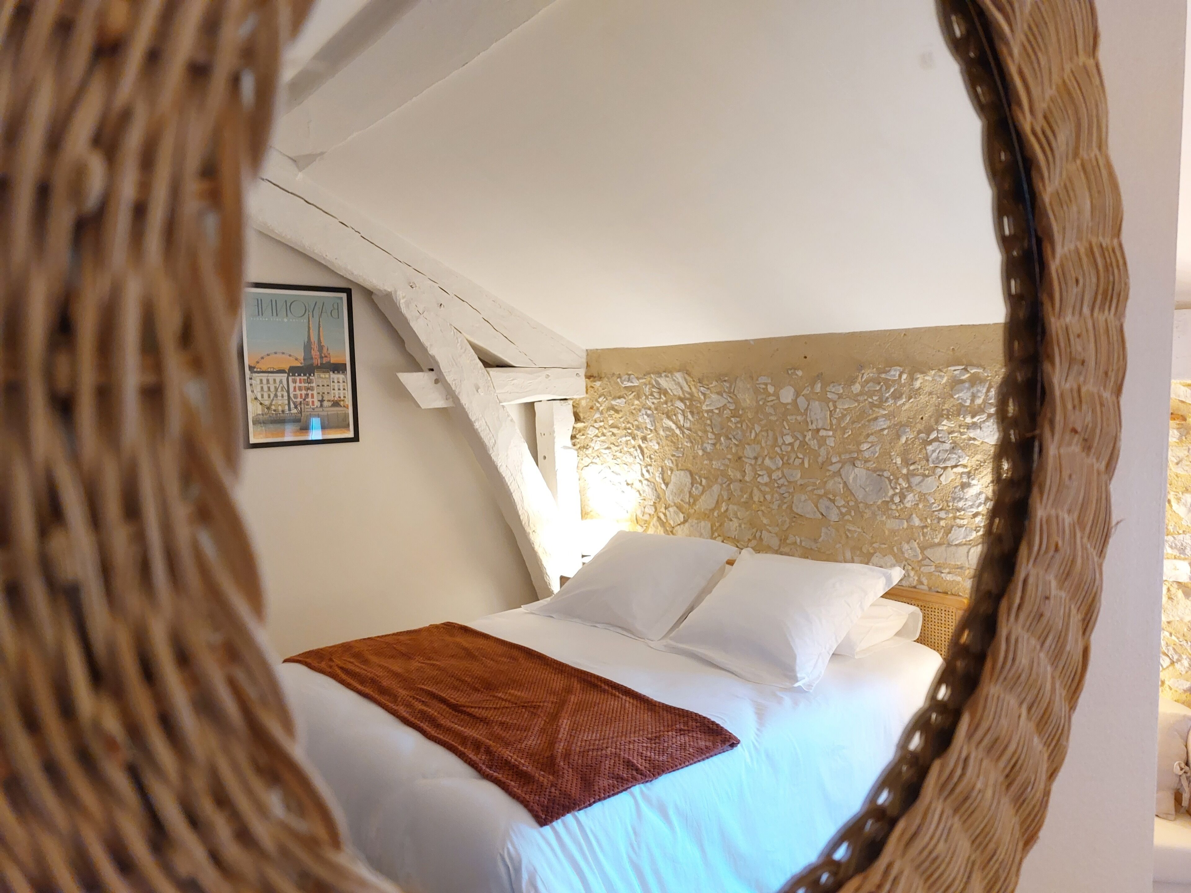 Een slaapkamer met een bed, wit beddengoed, een bruine plaid, een stenen muur en een lijst met een afbeelding aan de muur.