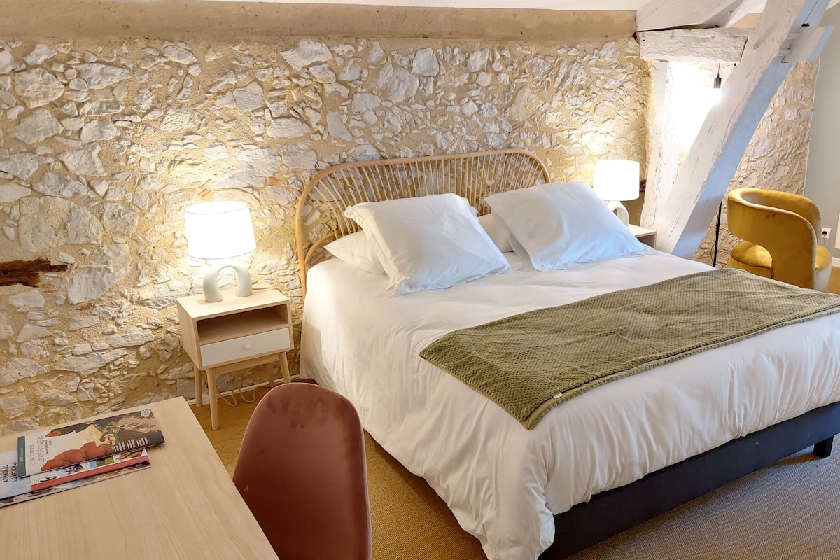 A bedroom with a stone wall, a bed with white bedding, a bedside table with a lamp, a chair, and a small table with a book.