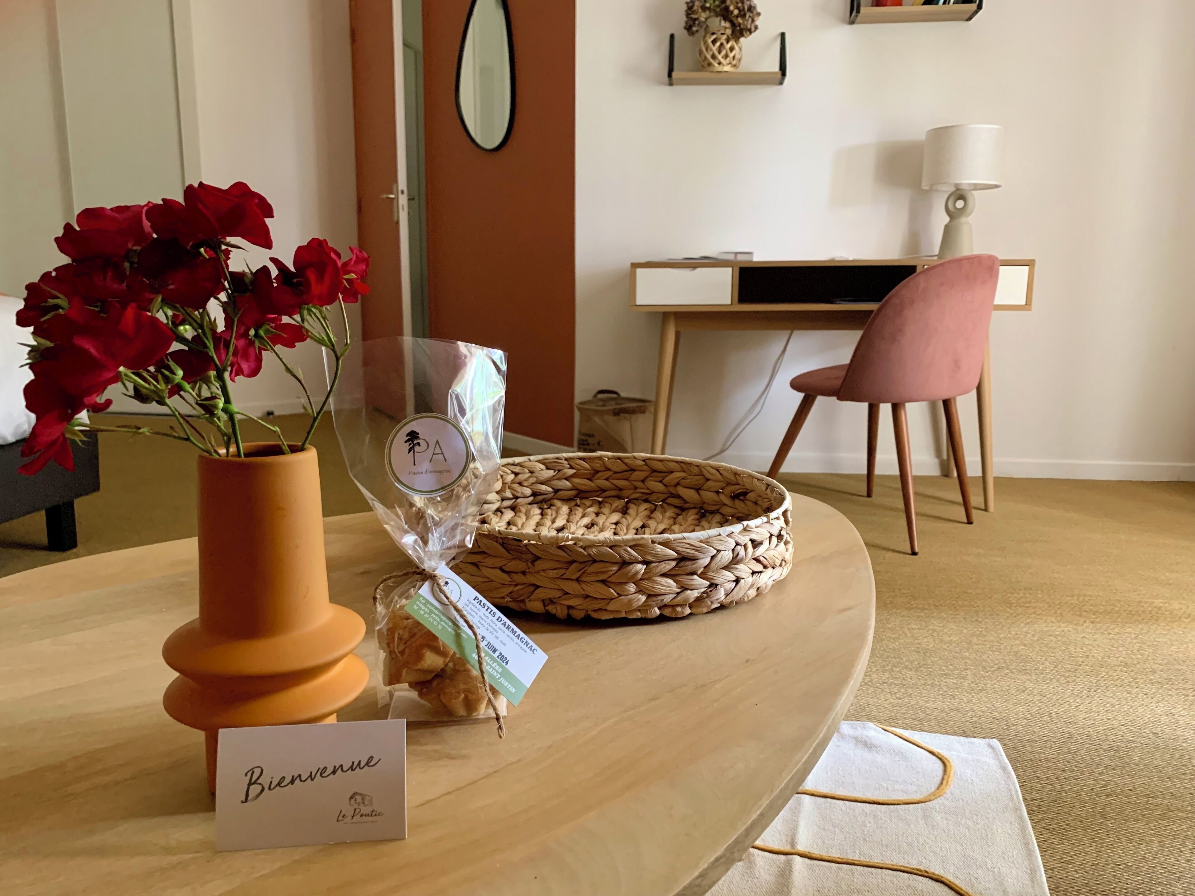 Una habitación con una mesa de madera, un jarrón con flores rojas, una cesta tejida y un letrero que dice “Bienvenue Le P'tic”.
