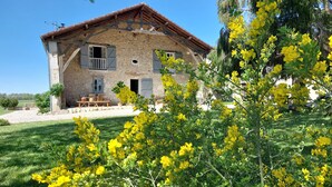 Una casa de piedra con balcón de madera, un área de comedor con mobiliario exterior y plantas florecientes amarillas en primer plano.