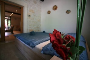 Quarto com parede de pedra, cama com roupa de cama azul e um vaso com rosas vermelhas.