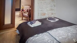 A hotel room with a large bed, a small table with a chair, and a stone wall.