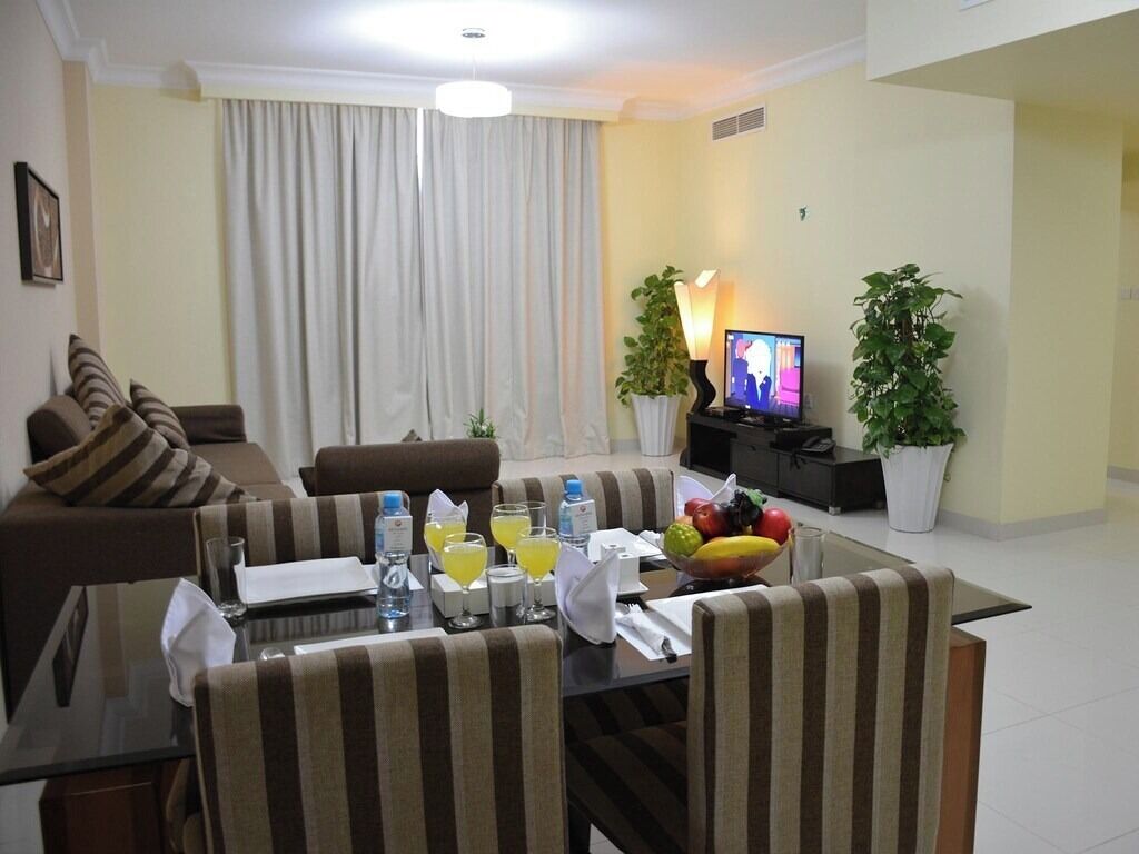 A modern living room with a dining area, featuring a sofa, striped chairs, a glass-top dining table with fruit and drinks, a TV, and potted plants.