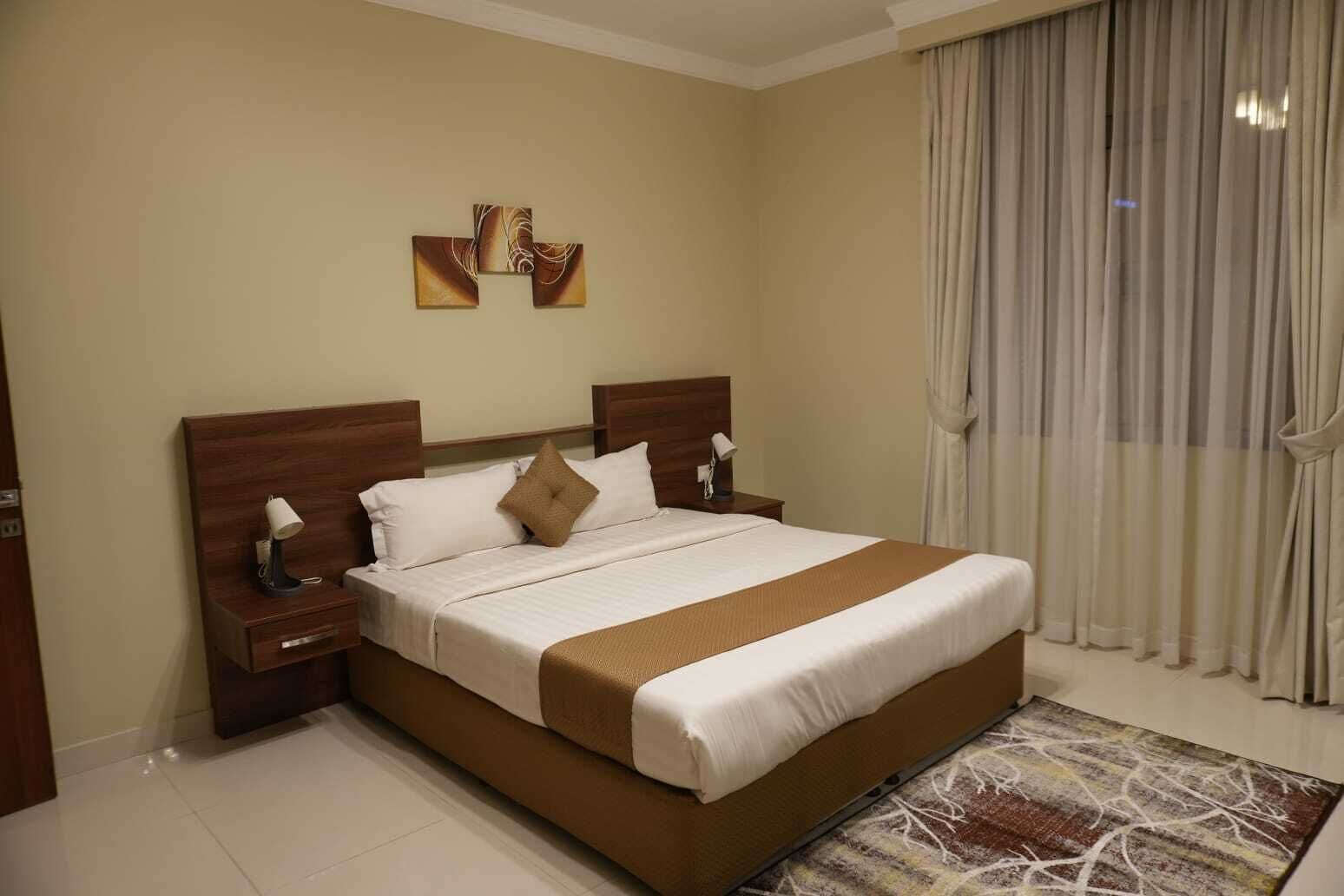 A hotel room with a large bed, wooden headboard, two bedside tables with lamps, a beige wall, and a window with curtains.