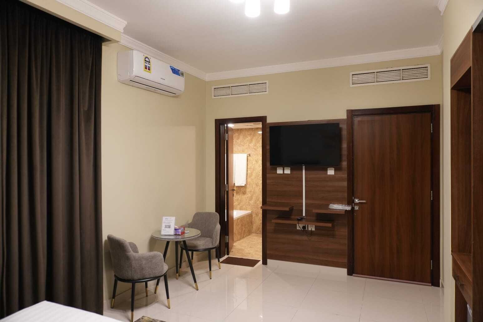 Une chambre d’hôtel avec un téléviseur à écran plat fixé sur un mur en bois, deux fauteuils gris, une petite table ronde, un lit blanc et une grande fenêtre avec des rideaux.