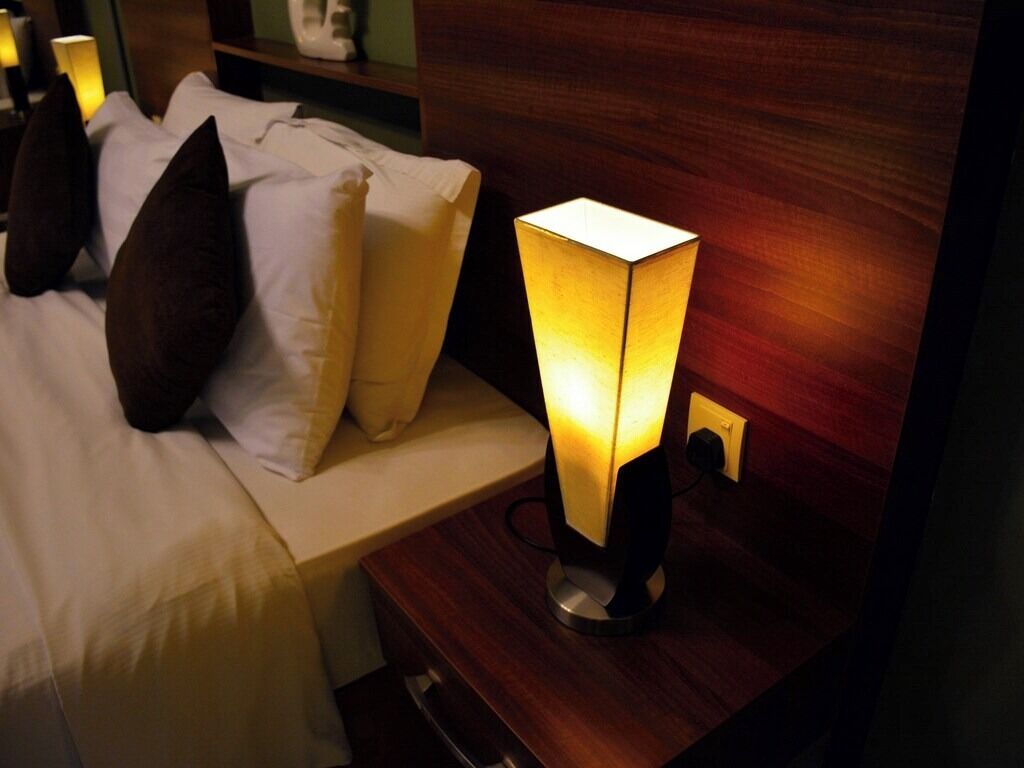 Una cama con sábanas blancas y almohadillas oscuras, una mesita de noche con una lámpara y un cabecero de madera.