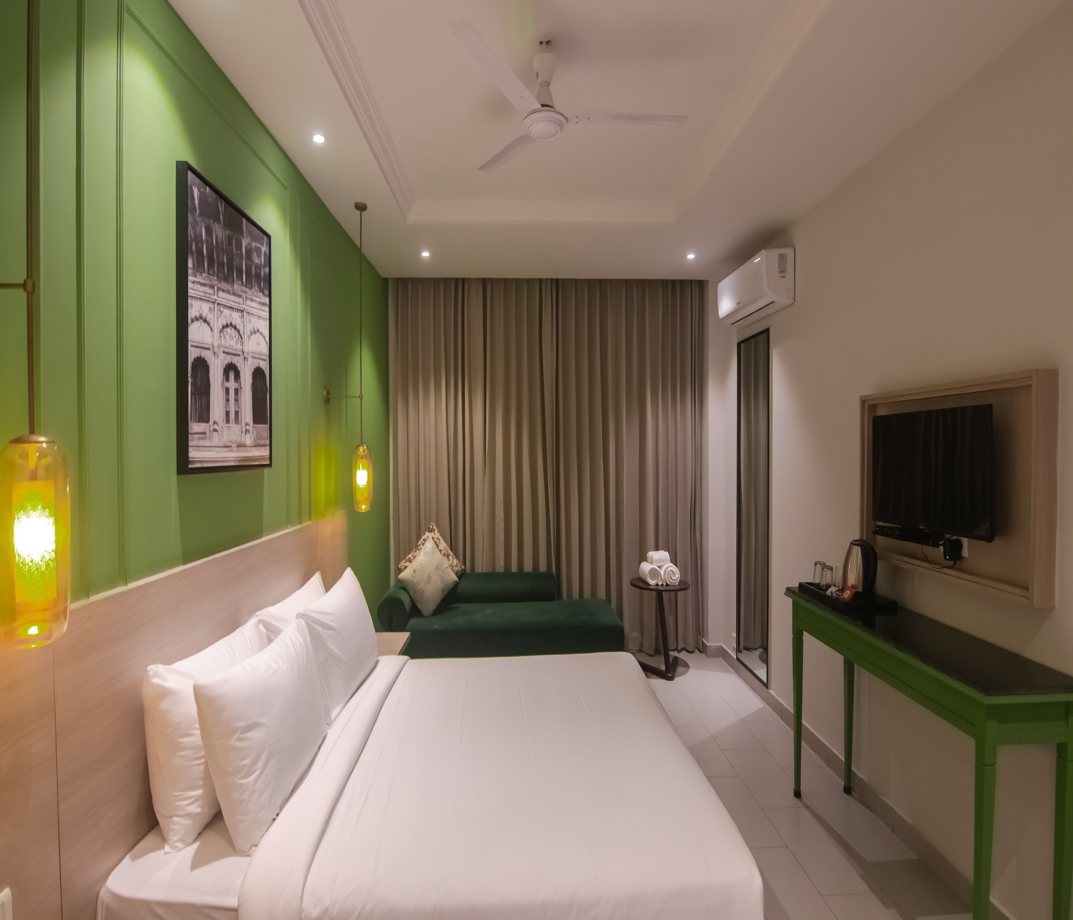 A hotel room with a bed, a green wall, a ceiling fan, a mounted TV, and a window with curtains.