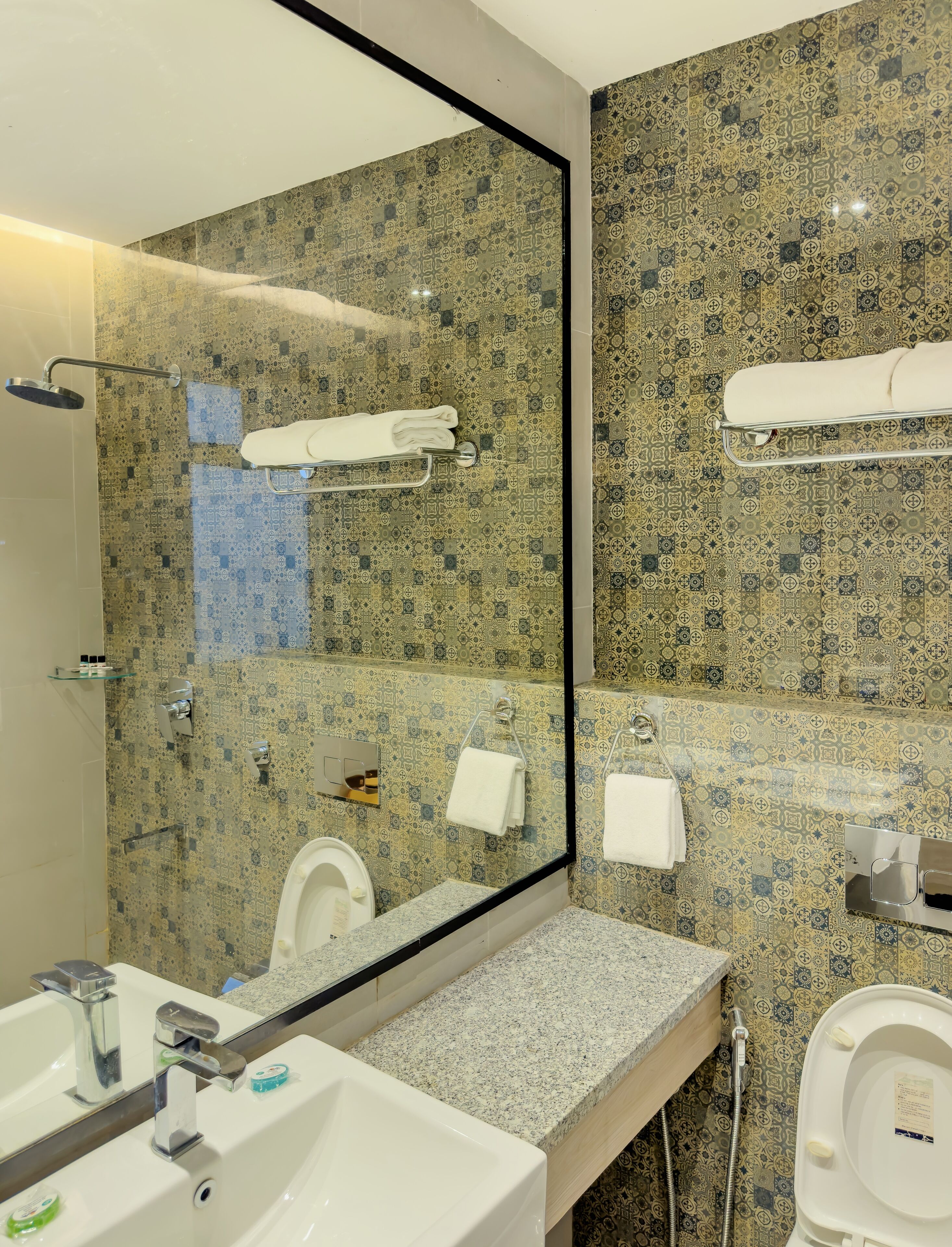 A bathroom with a patterned tile wall, a large mirror, a sink, a toilet, and a shower area.