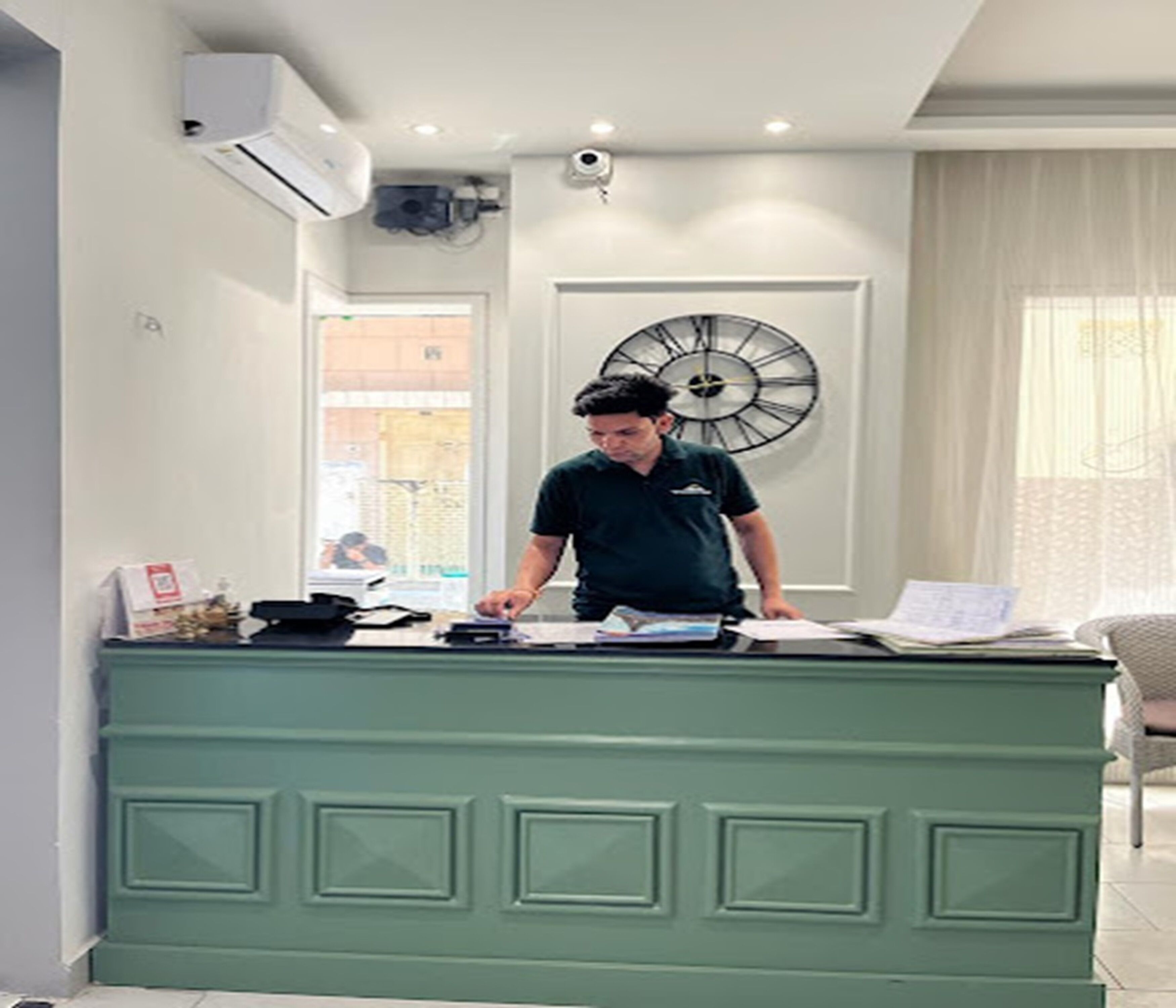 A person at a green reception desk with papers, a clock, and a window in the background.