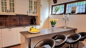 A modern kitchen with white cabinets, a central island with a fruit bowl, and a window with a view of greenery. - TG Oasis Örgryte Stugor & Rum (Gothenburg)