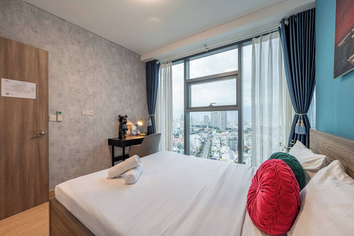 A hotel room with a large bed, a desk with a lamp, a view of the city, and a wooden wardrobe.