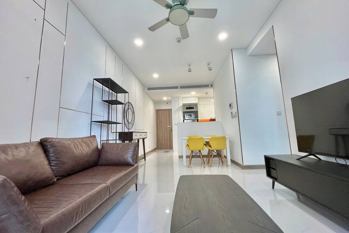 A modern living room with a brown leather sofa, a grey coffee table, and a TV on a black stand. There is a ceiling fan, recessed lighting, and a kitchen area in the background.