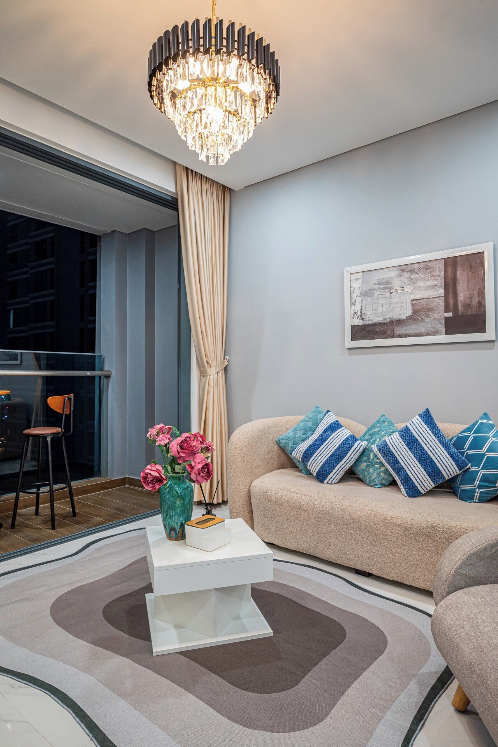 A modern living room with a beige sofa, blue and white striped pillows, a white coffee table with a vase of pink flowers, a large chandelier, and a framed abstract painting on the wall.