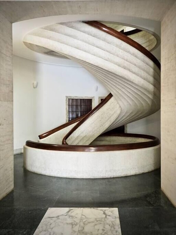 A spiral staircase with a wooden handrail and white steps, located in a hallway with a marble floor.