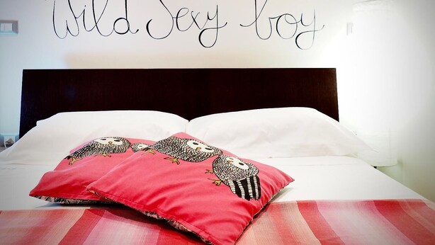 Une chambre à coucher avec un lit, des oreillers, un mur orné d’un motif à rayures rouges et blanches, et un autre mur sur lequel on peut lire le texte suivant : « Wild Sexy boy ».