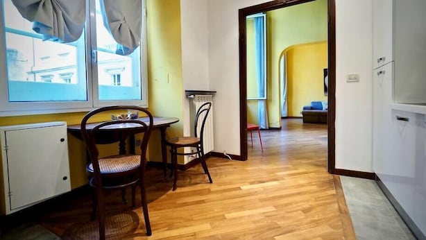 Een kamer met houten vloer, een eettafel met stoelen, een raam met gordijnen en een deur naar een andere kamer.