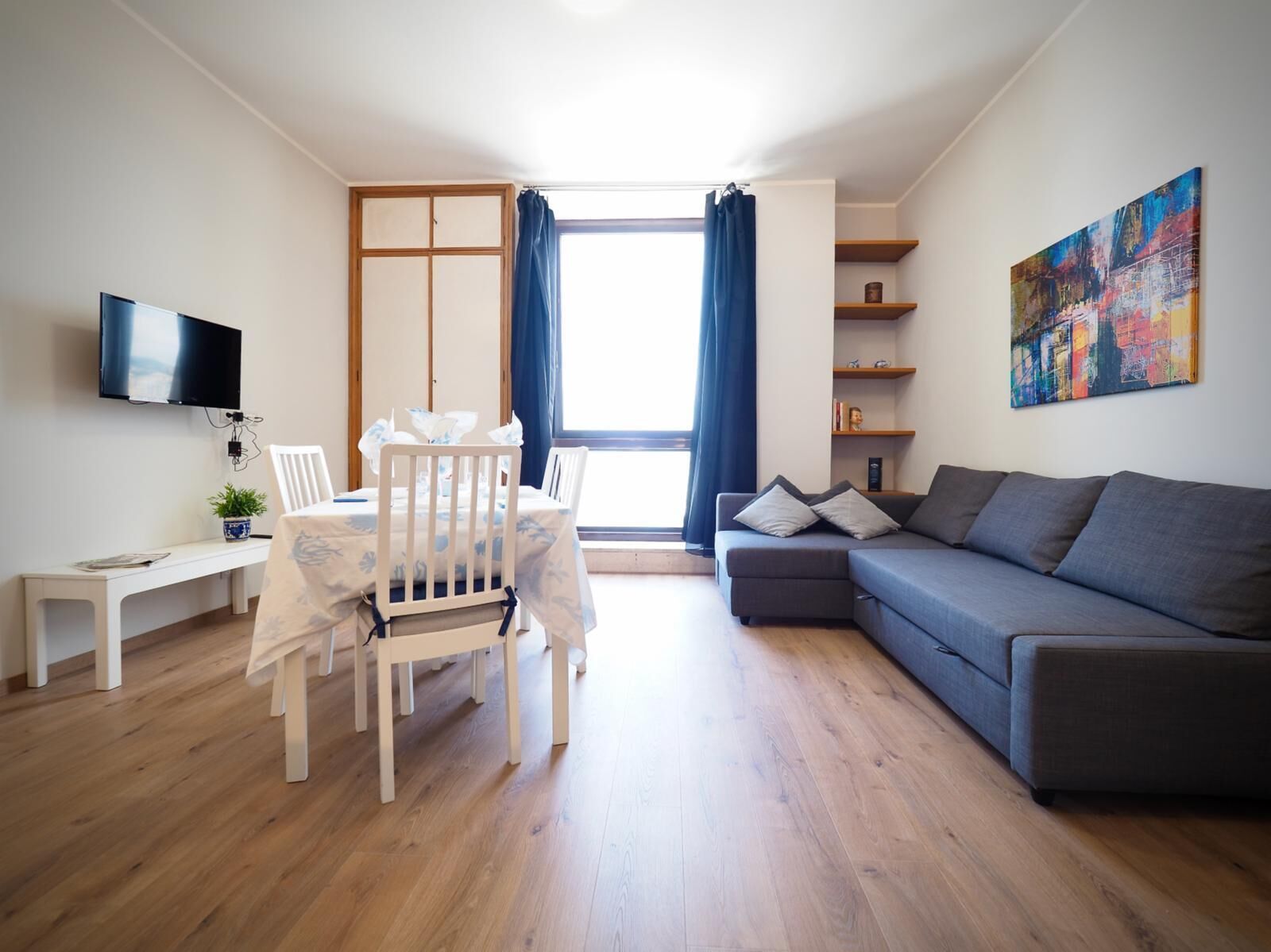 Un salon moderne avec un plancher en bois, un canapé gris, une table à manger blanche avec des chaises, un téléviseur fixé au mur et une grande fenêtre avec des rideaux.