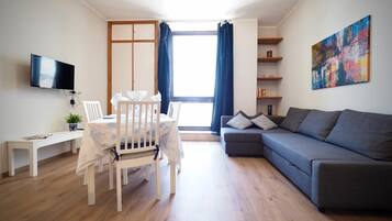 Un salon moderne avec un plancher en bois, un canapé gris, une table à manger blanche avec des chaises, un téléviseur fixé au mur et une grande fenêtre avec des rideaux.