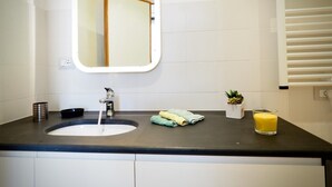 Un baño moderno con una encimera negra, un lavabo con grifo, un espejo, una planta pequeña, una toalla y un vaso de zumo de naranja.