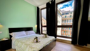 Un dormitorio con una cama grande, dos mesitas de noche, una ventana con cortinas y vistas a edificios.