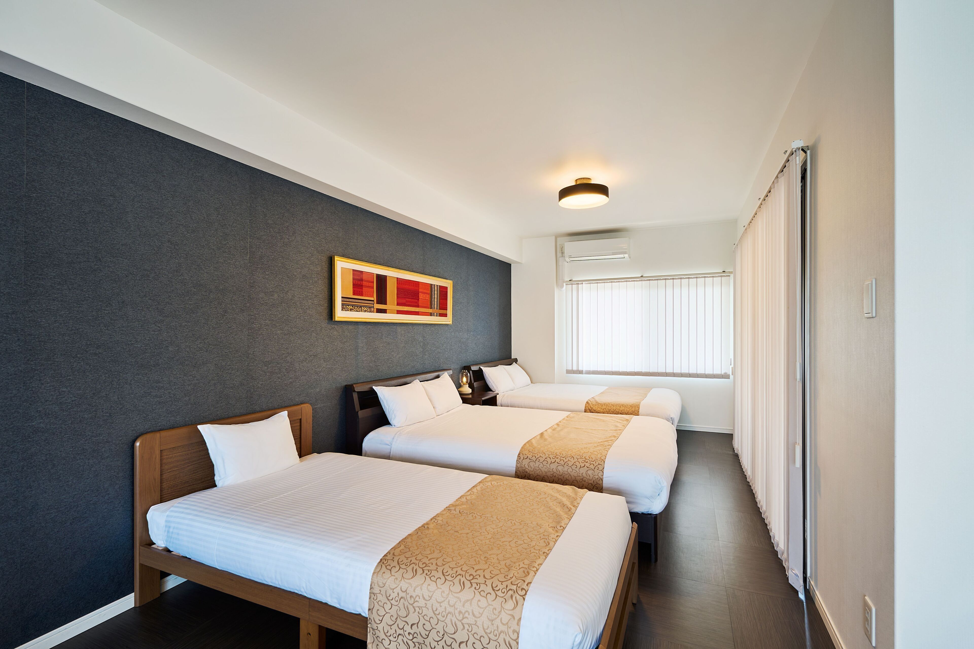A hotel room with three beds, a painting on the wall, and a window with blinds.