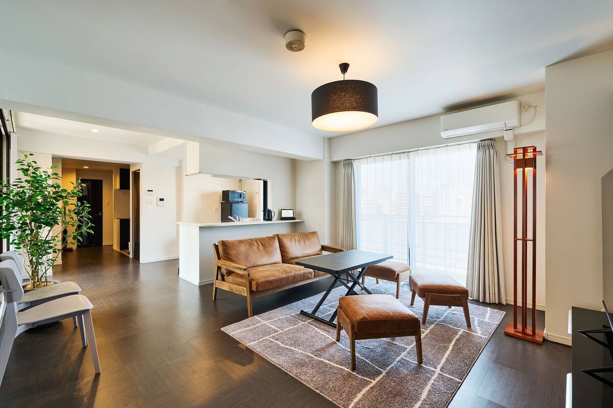 A modern living room with a sofa, coffee table, and ottoman, a dining area with chairs, a large window with curtains, and a floor lamp.