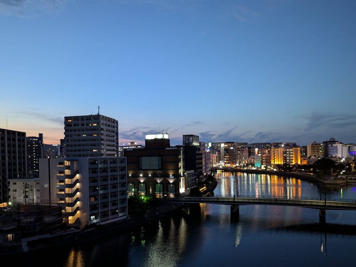 A cityscape with modern buildings along a river, a bridge crossing the water, and illuminated windows suggesting nighttime.