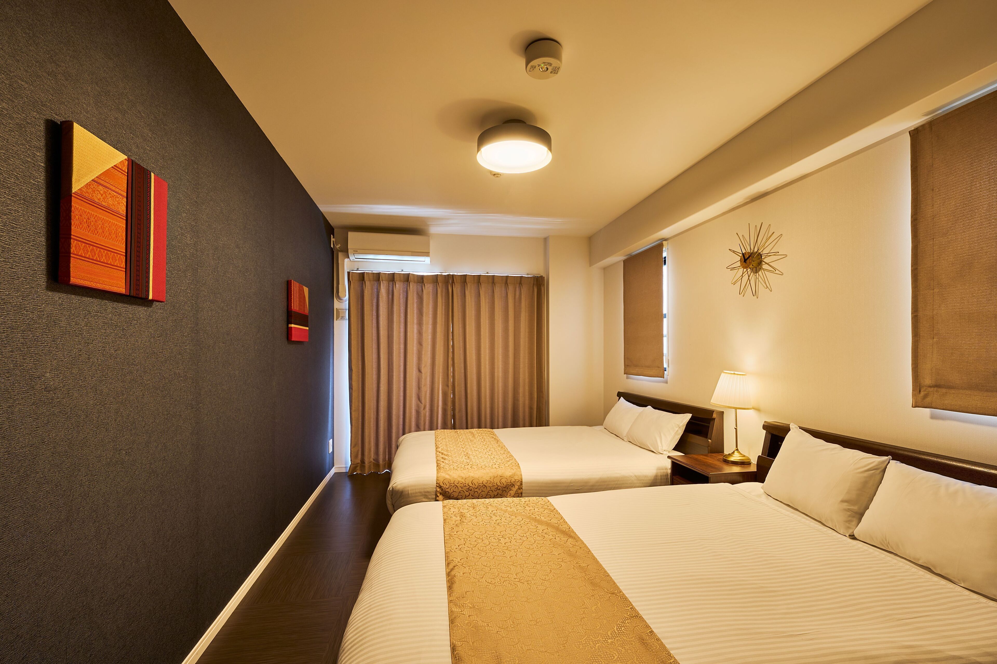 A hotel room with two beds, a nightstand with a lamp, a wall clock, and a window with curtains.