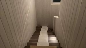 Eine enge Treppe mit abwechselnd dunklen und hellen Stufen, ein kleines Fenster am Obergeschoss und eine Leuchte darüber.