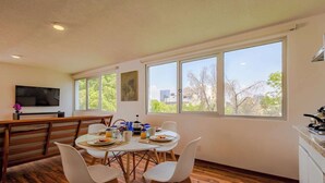 A dining area with a table set for four, a television mounted on the wall, and large windows offering a view of trees and buildings.