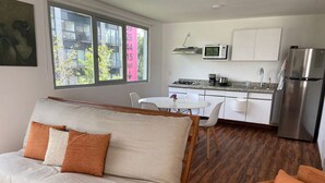 A modern living room with a sofa and armchair, a kitchen with white cabinets and stainless steel appliances, and a large window with a view of buildings and trees.