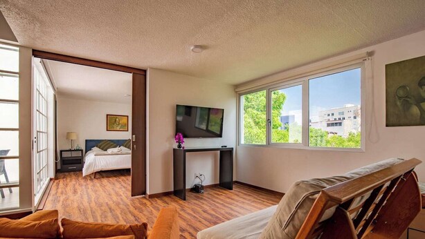 A modern hotel room with a bed, a desk with a TV, a sofa, and a large window offering a city view.