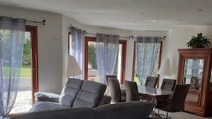 Ein geräumiges Wohnzimmer mit grauem Sofa, einem Esstisch mit Stühlen und großen Fenstern mit durchsichtigen Vorhängen.