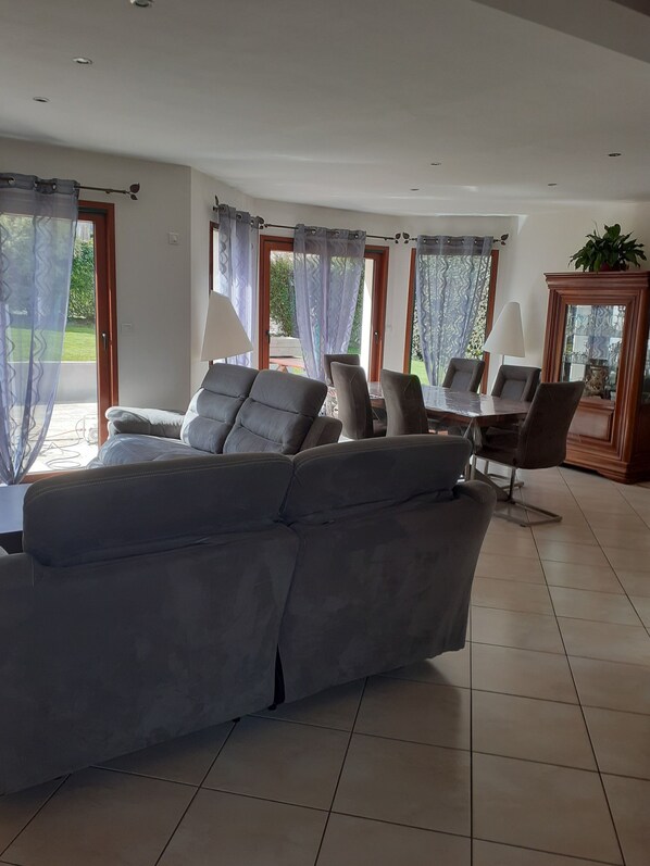 A spacious living room with a grey sofa, a dining table with chairs, and large windows with sheer curtains.