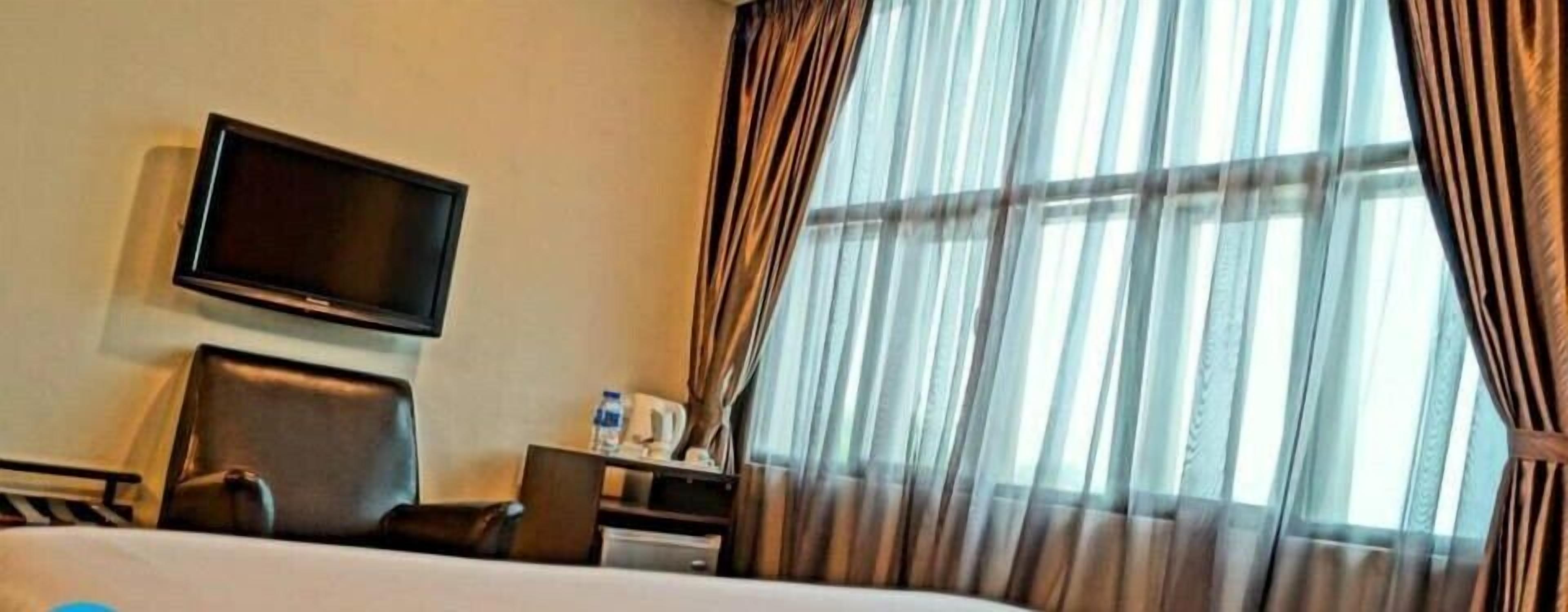 Habitación de hotel con cama, televisor de pantalla plana montado en la pared, escritorio con silla y ventana con cortinas traslúcidas.