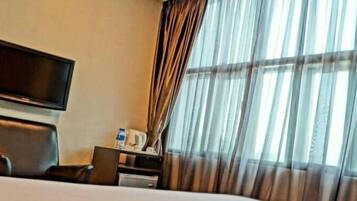 A hotel room with a bed, a flat-screen TV mounted on the wall, a desk with a chair, and a window with sheer curtains.