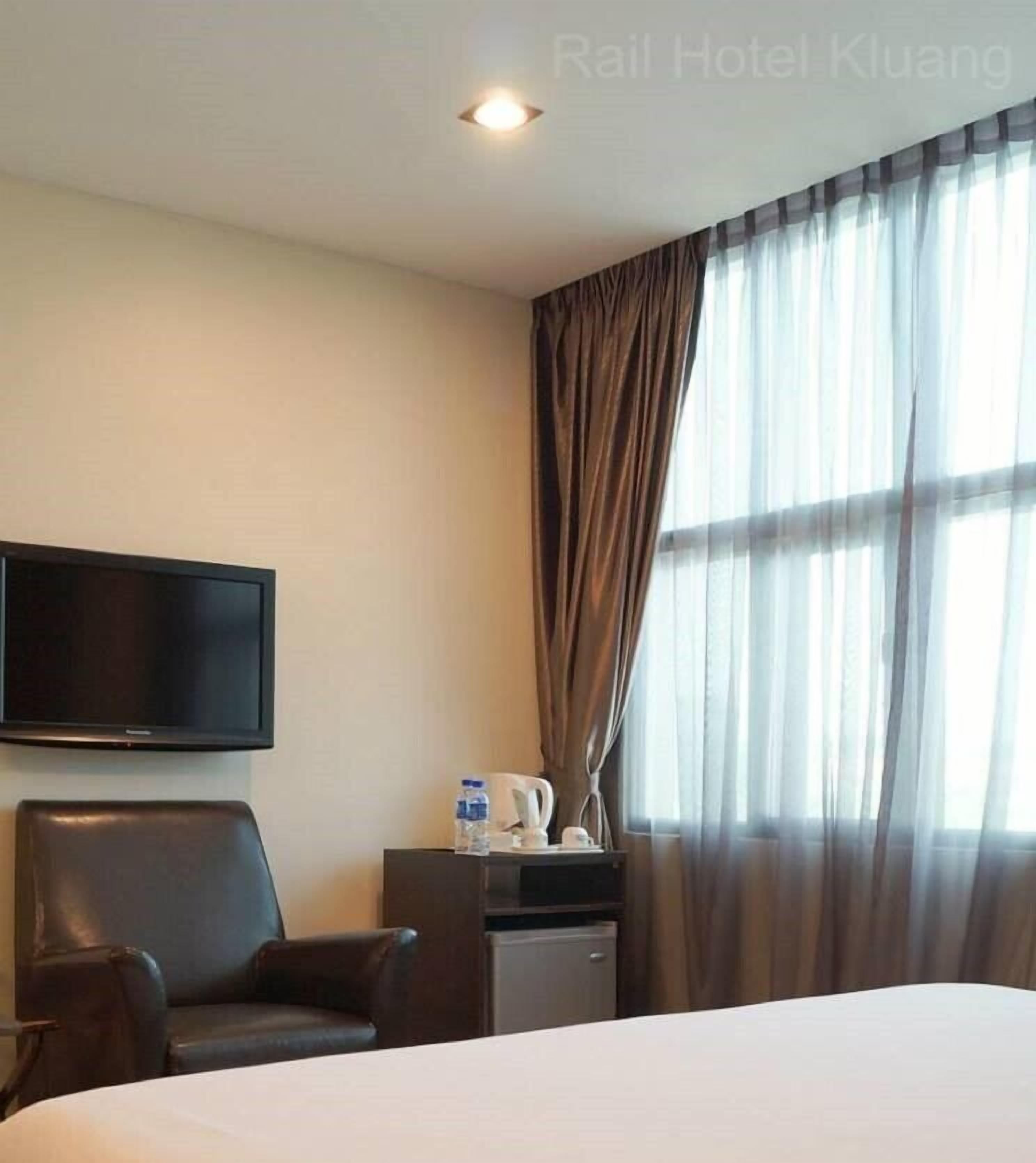 A hotel room with a bed, a flat-screen TV mounted on the wall, a desk with a kettle, a chair, and a window with curtains.