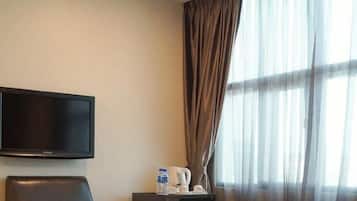 A hotel room with a bed, a flat-screen TV mounted on the wall, a desk with a kettle, a chair, and a window with curtains.