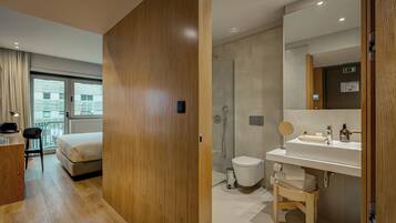Una habitación de hotel moderna con piso de madera, una cama, un escritorio con una silla, un baño con ducha, un lavamanos y un espejo.