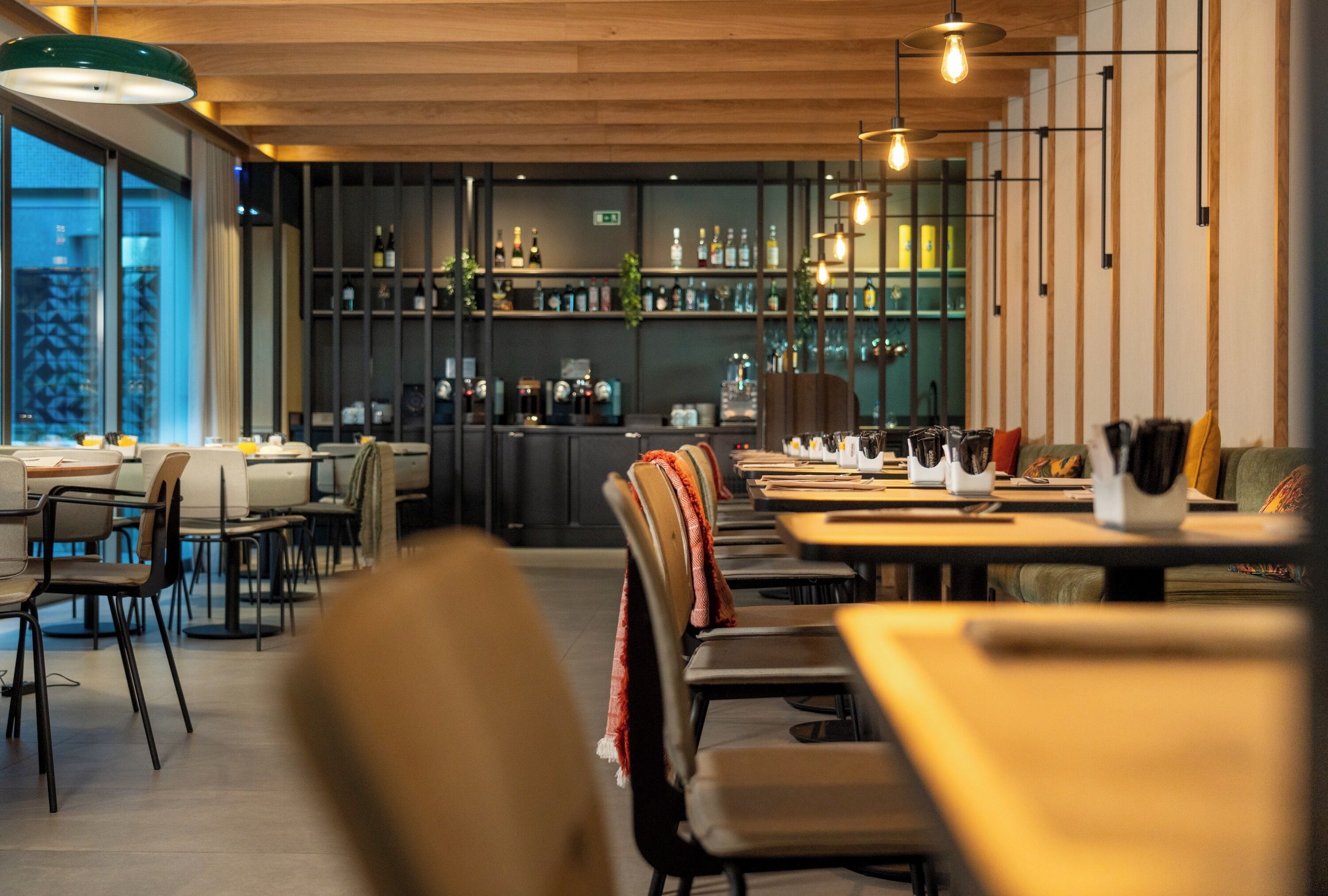 A modern restaurant interior with wooden tables and chairs, a bar area with shelves of bottles, and pendant lighting.