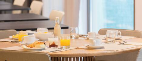 Una mesa de desayuno dispuesta con platos, tazones, vasos y una botella, en una habitación con sillas y vistas a un edificio a través de una ventana.
