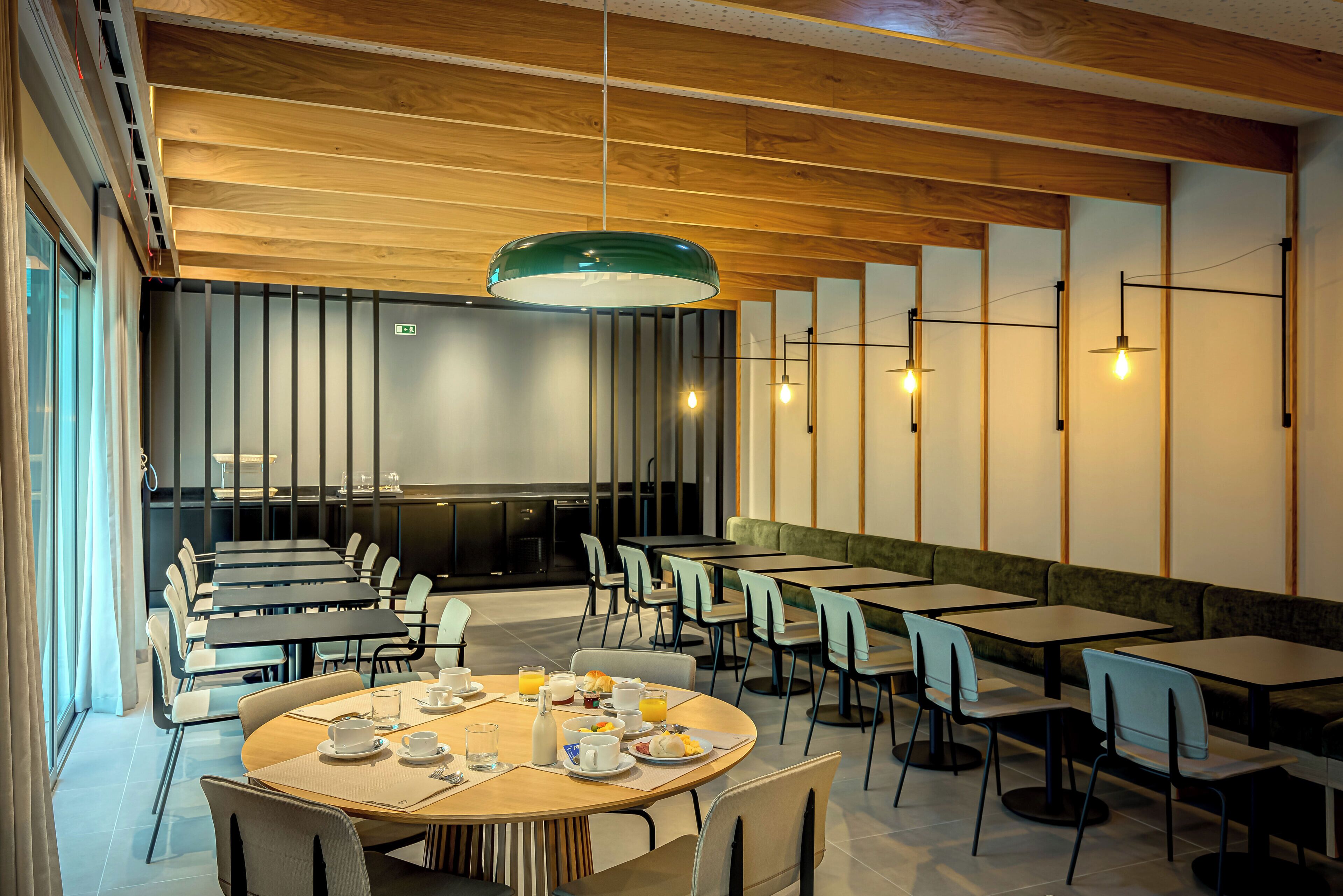 A modern dining area with multiple tables and chairs, a large green pendant light, and a kitchen area in the background.
