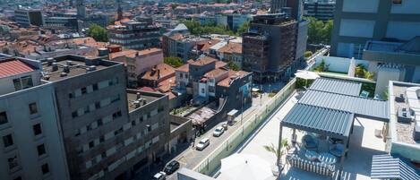 Un edificio de gran altura con varios pisos, una terraza en la azotea con asientos y vistas de un paisaje urbano con diferentes edificios y una torre distante.