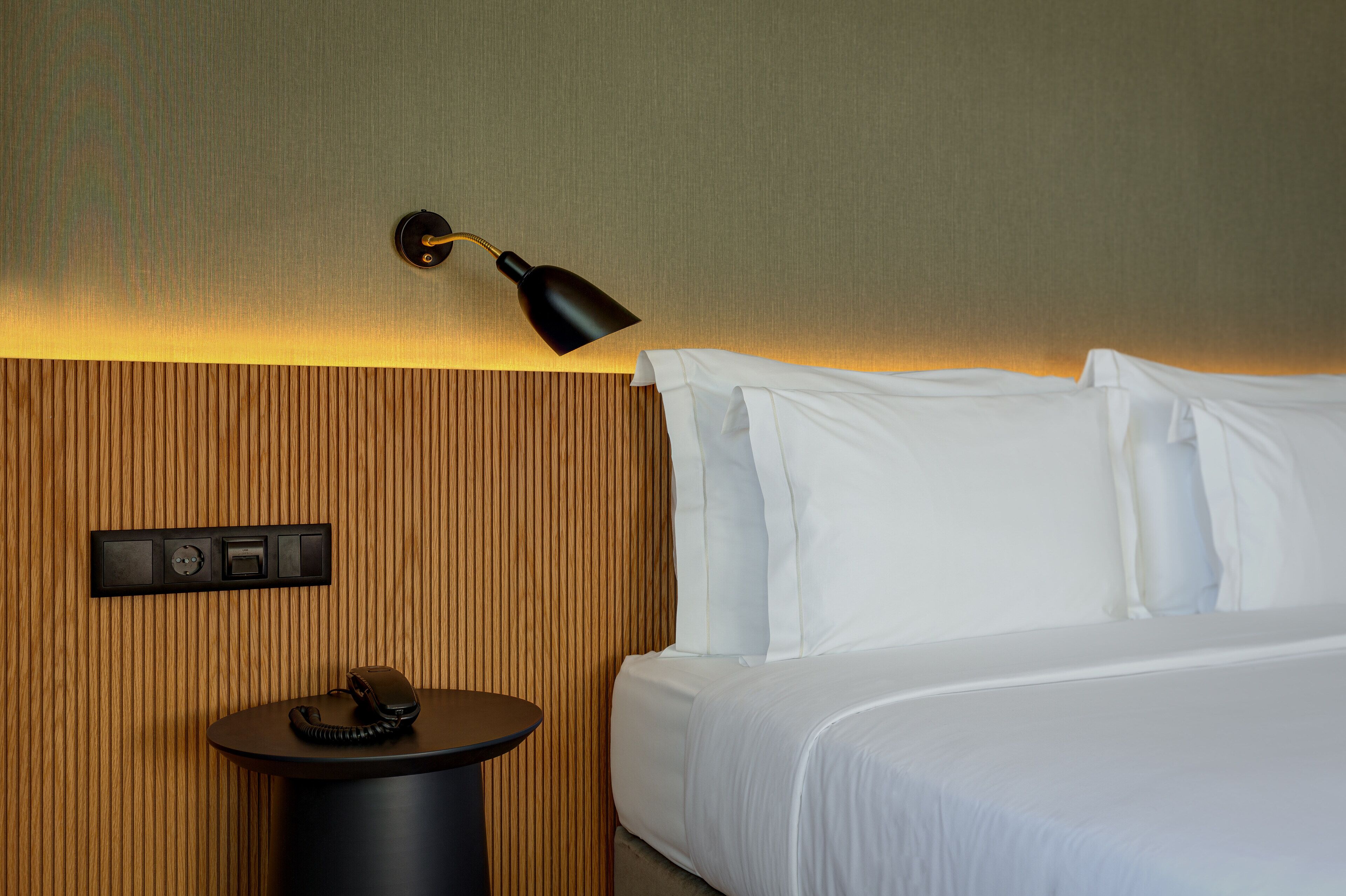 A hotel room with a bed, a bedside table with a telephone, a wall-mounted light, and a control panel.