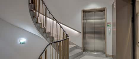 Un ascensor moderno con detalles metálicos, una escalera curva con barandillas de madera y un letrero de salida de emergencia instalado en la pared.