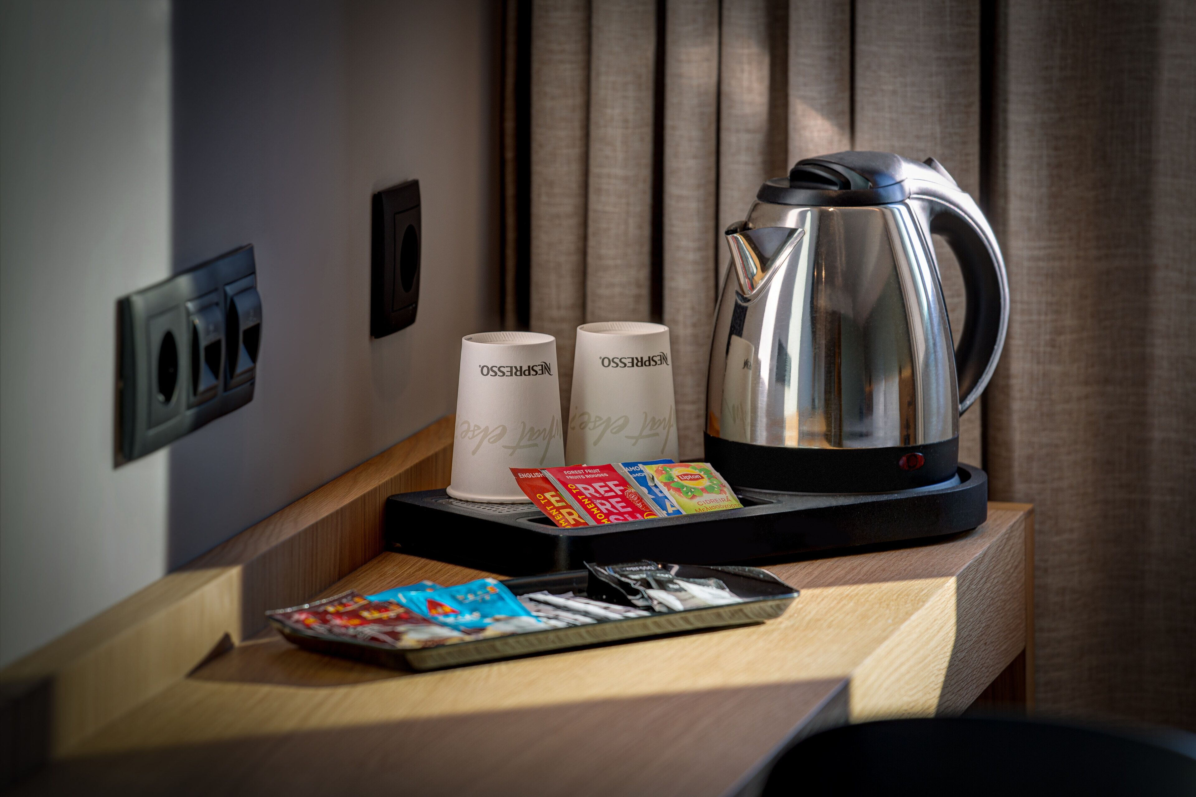 A hotel room with a wooden side table holding a coffee maker, two Nespresso cups, a tray with cigarettes and a magazine.